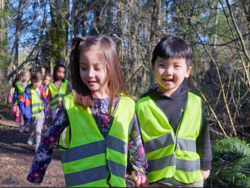 2 students holding hands wearing safety vests and walking in the woods with their class