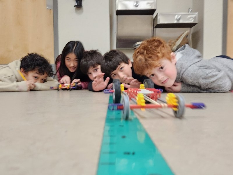 students down on the floor testing a car made from pencils, rubber bands and other materials