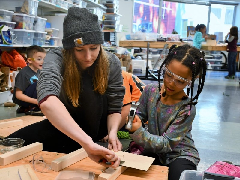 young student wearing safety glasses as a teacher helps them use a hammer for a project in the makerspace