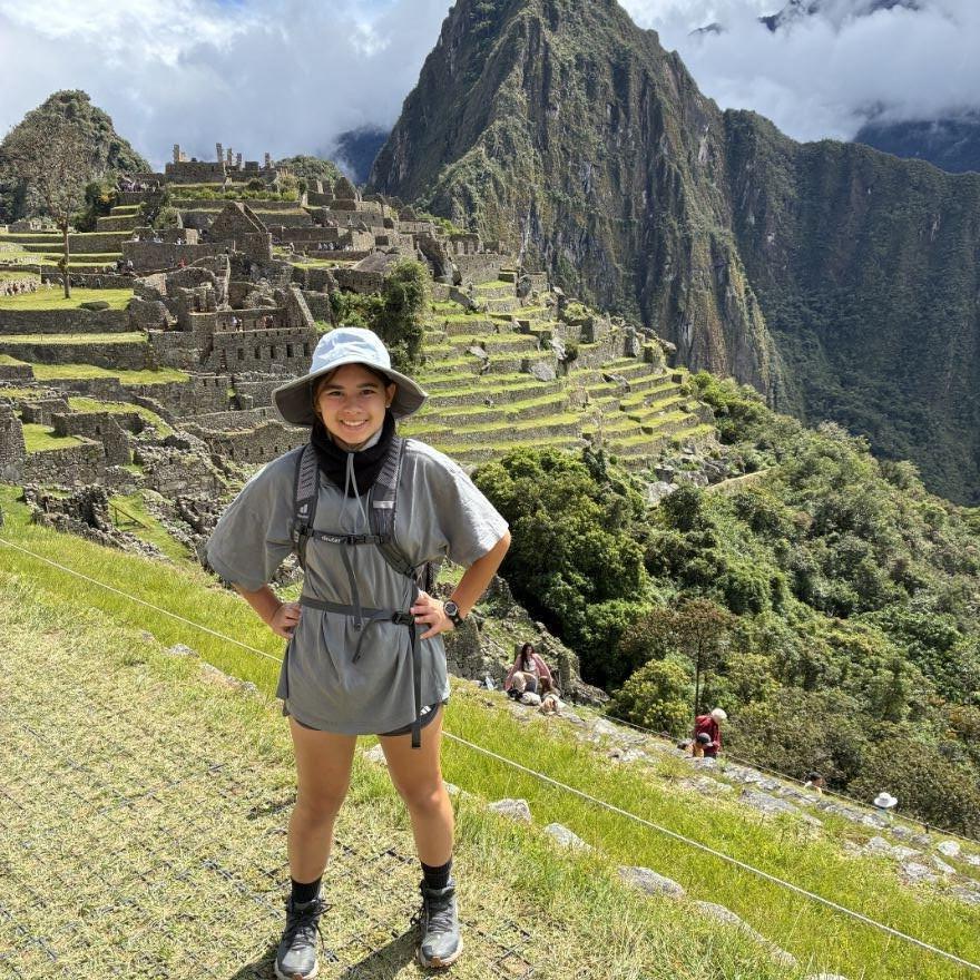 A student in hiking gear, smiling and posing proudly on a trail above Machu Picchu. Terraced ruins and grass are behind them.