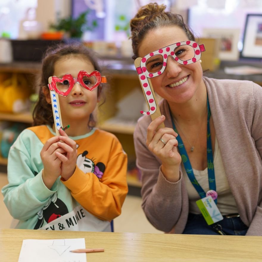 kindergarten student and teacher smiling and peering through pretend glasses decorated with hearts and polka dots