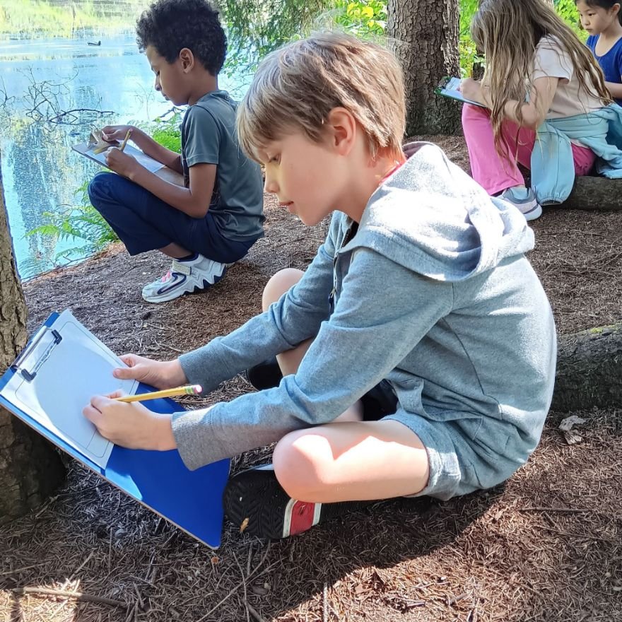 young students kneeling beside a pond, writing and drawing on clipboards