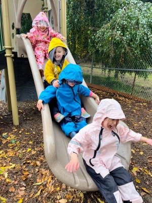 several young children sliding down a slide together, laughing and all wearing brightly colored, hooded rain suits